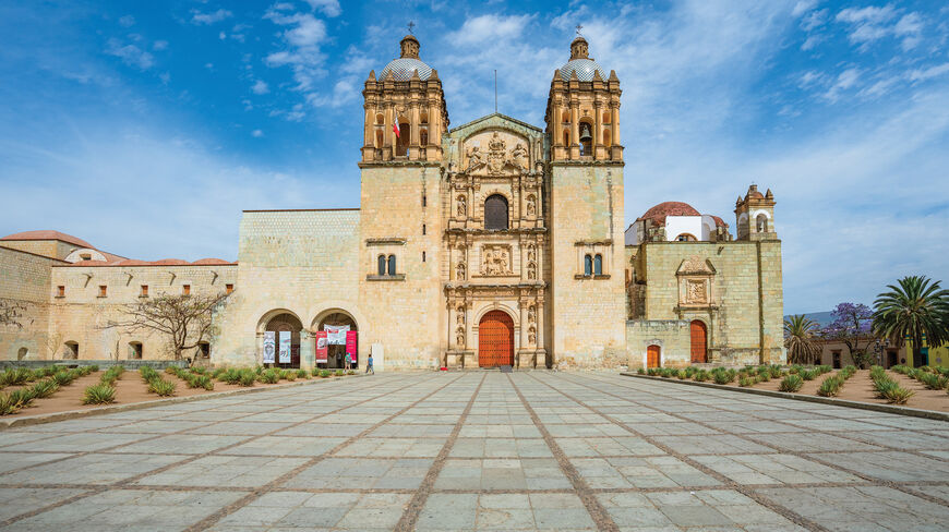 The ornate stone facade and bell towers of the Templo de Santo Domingo de Guzmán in Oaxaca, Mexico, seen from its large, stone-paved plaza.