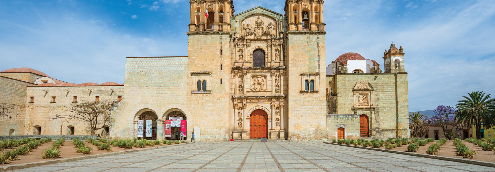 The ornate stone facade and bell towers of the Templo de Santo Domingo de Guzmán in Oaxaca, Mexico, seen from its large, stone-paved plaza.