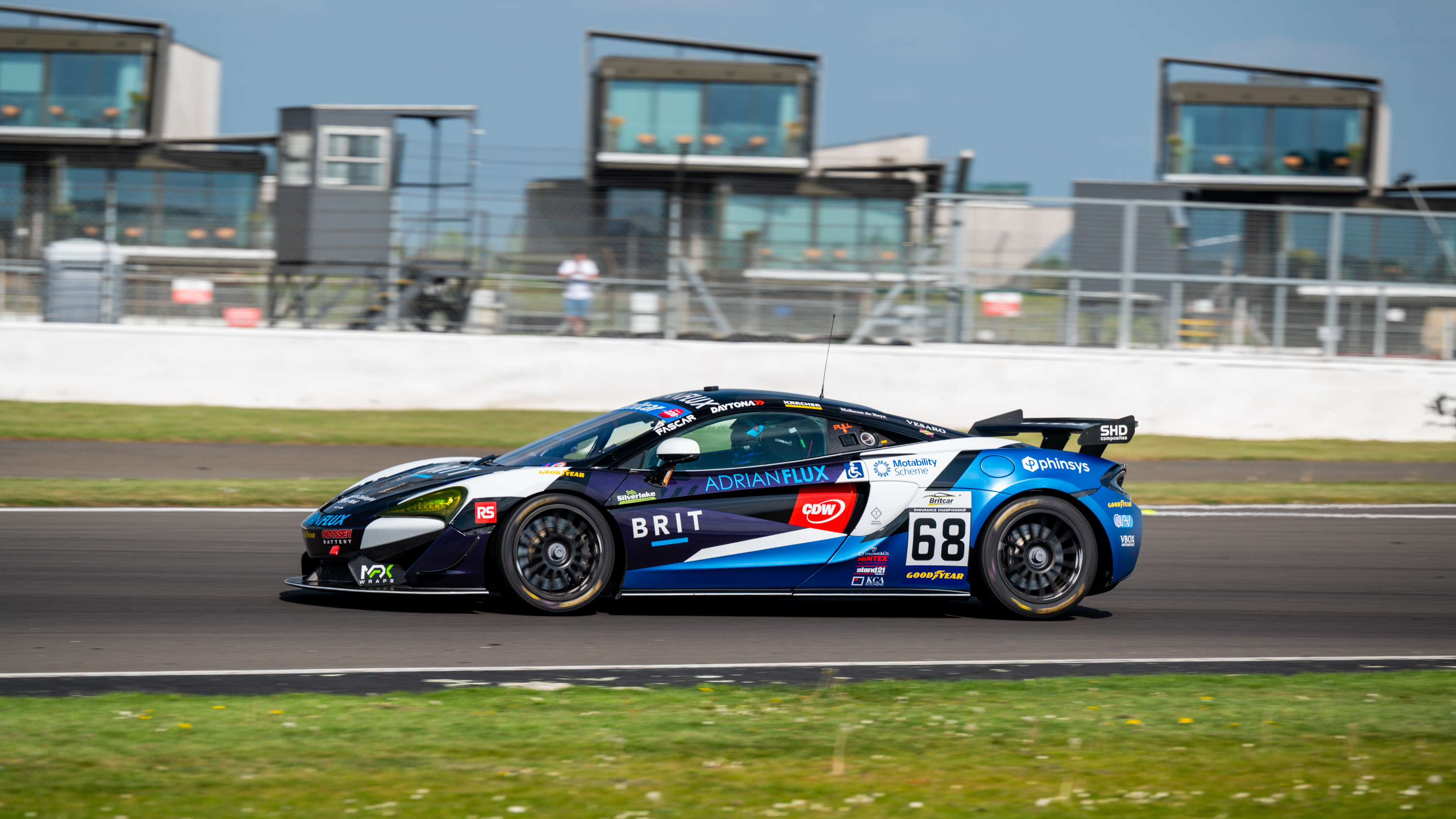 A blue and white Team BRIT race car speeds along a racetrack, displaying sponsor logos including Motability Scheme, with modern trackside buildings in the background.