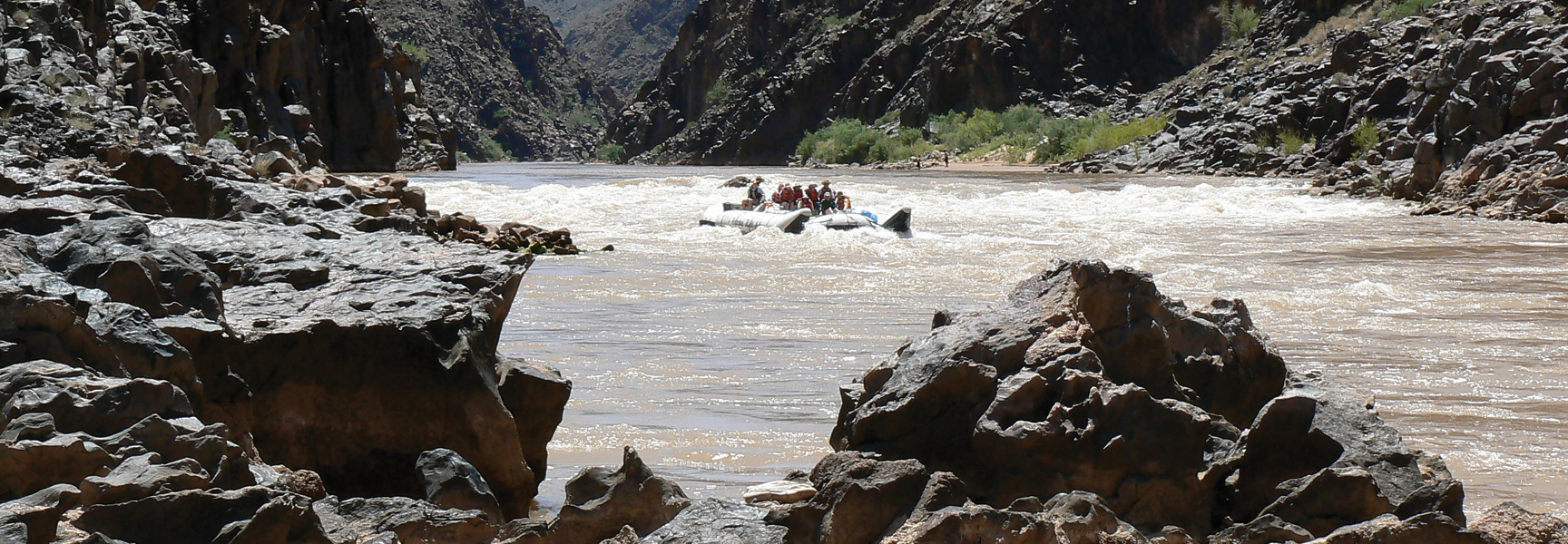 A group of people in a large raft navigate white water rapids through the steep, rocky canyon walls of the Grand Canyon in Arizona.
