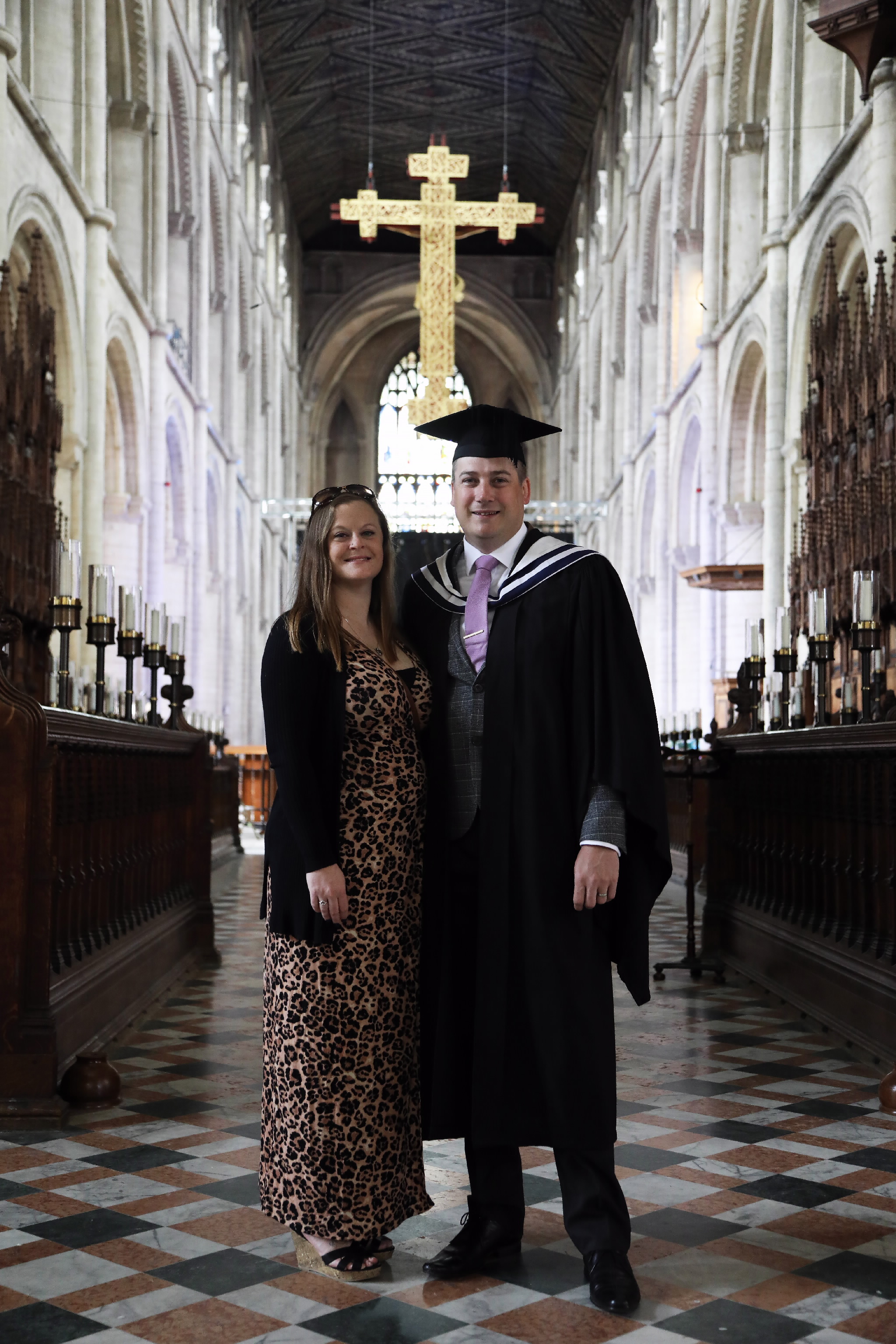 A graduate and a woman posing beneath a large cross