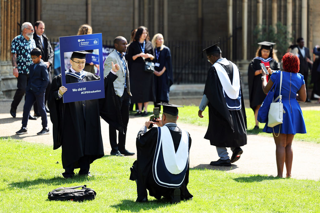 A graduate holding a 'We did it!' CIPS photo frame while another person takes a picture outdoors