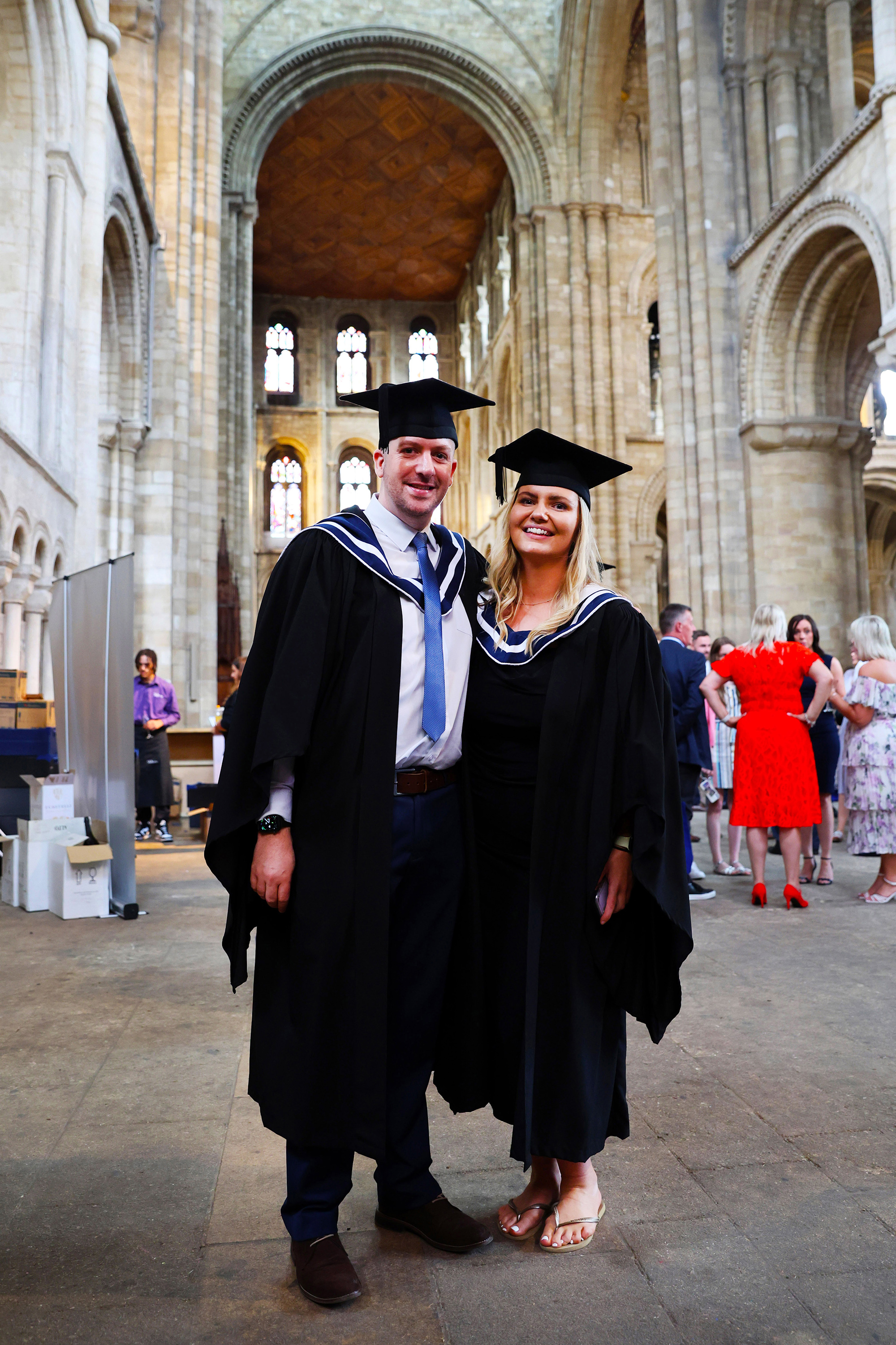 A happy couple in full black academic gowns and mortarboards posing inside the ornate, stone interior of a large cathedral