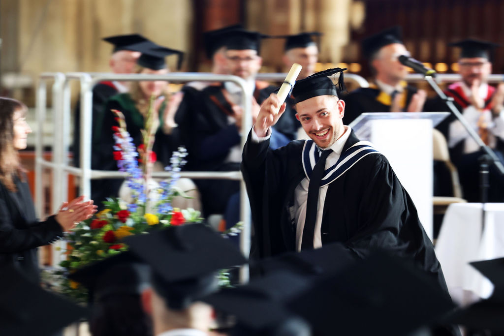 A male graduate on stage smiles and raises his diploma scroll to a clapping audience