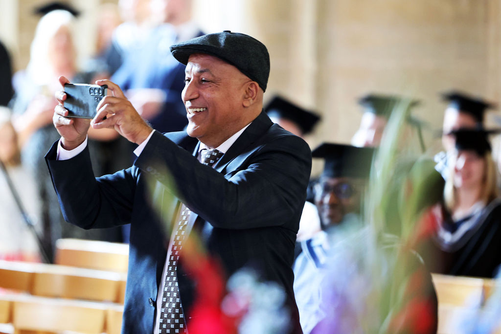 A smiling man in a suit and flat cap taking a photo with his phone during the graduation ceremony