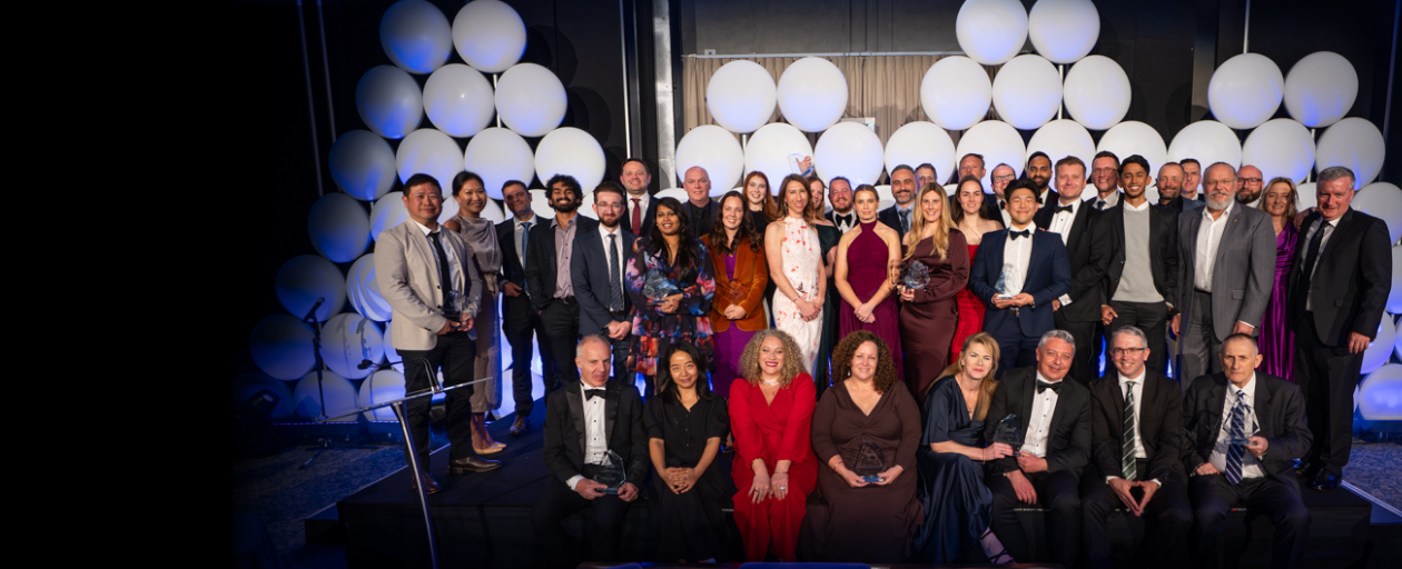 Group photo of award winners and presenters at a formal ceremony, with approximately 40 people in tuxedos, suits, and evening gowns, posing on a stage in front of a backdrop of white balloons and holding awards.