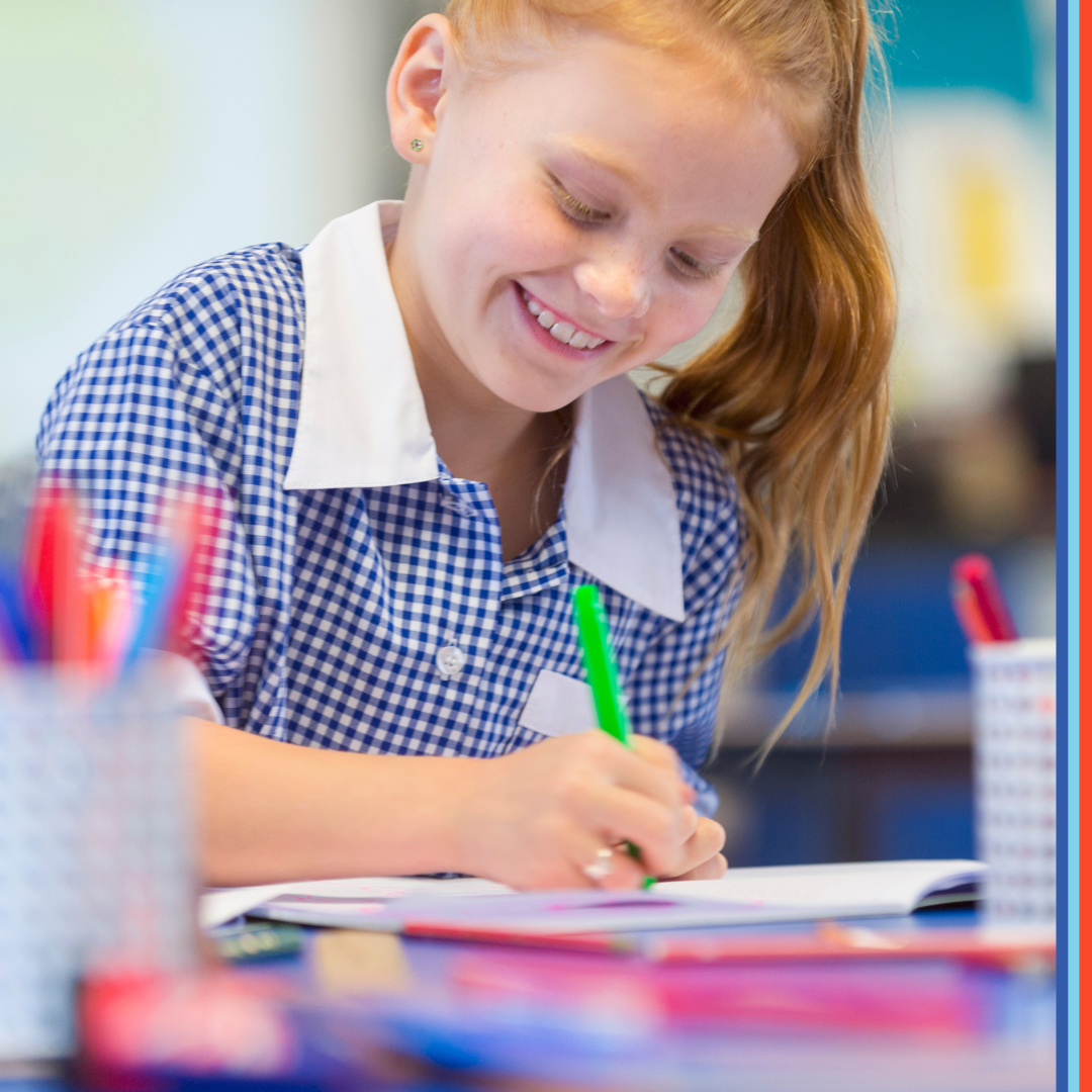 A schoolgirl is working at a desk. She is wearing a school uniform. She is writing or drawing with a felt pen. She is smiling and happy having fun.