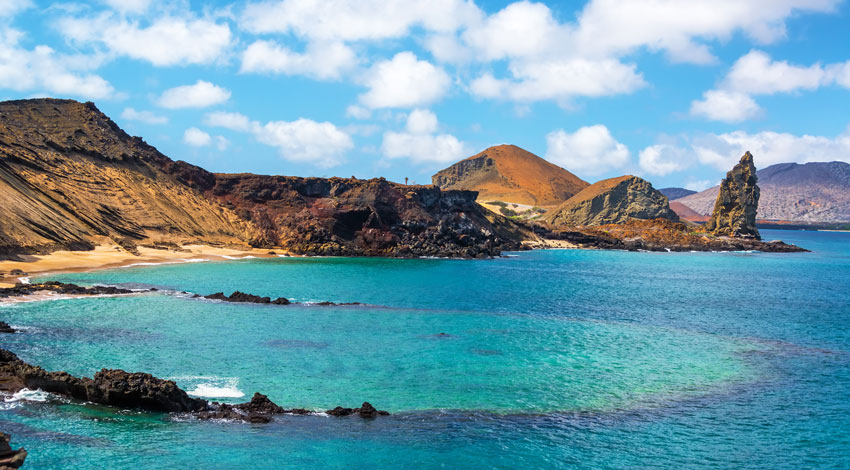 A stunning view of the Galapagos Islands featuring volcanic hills surrounding a turquoise bay with a sandy beach and a distinctive tall rock formation.