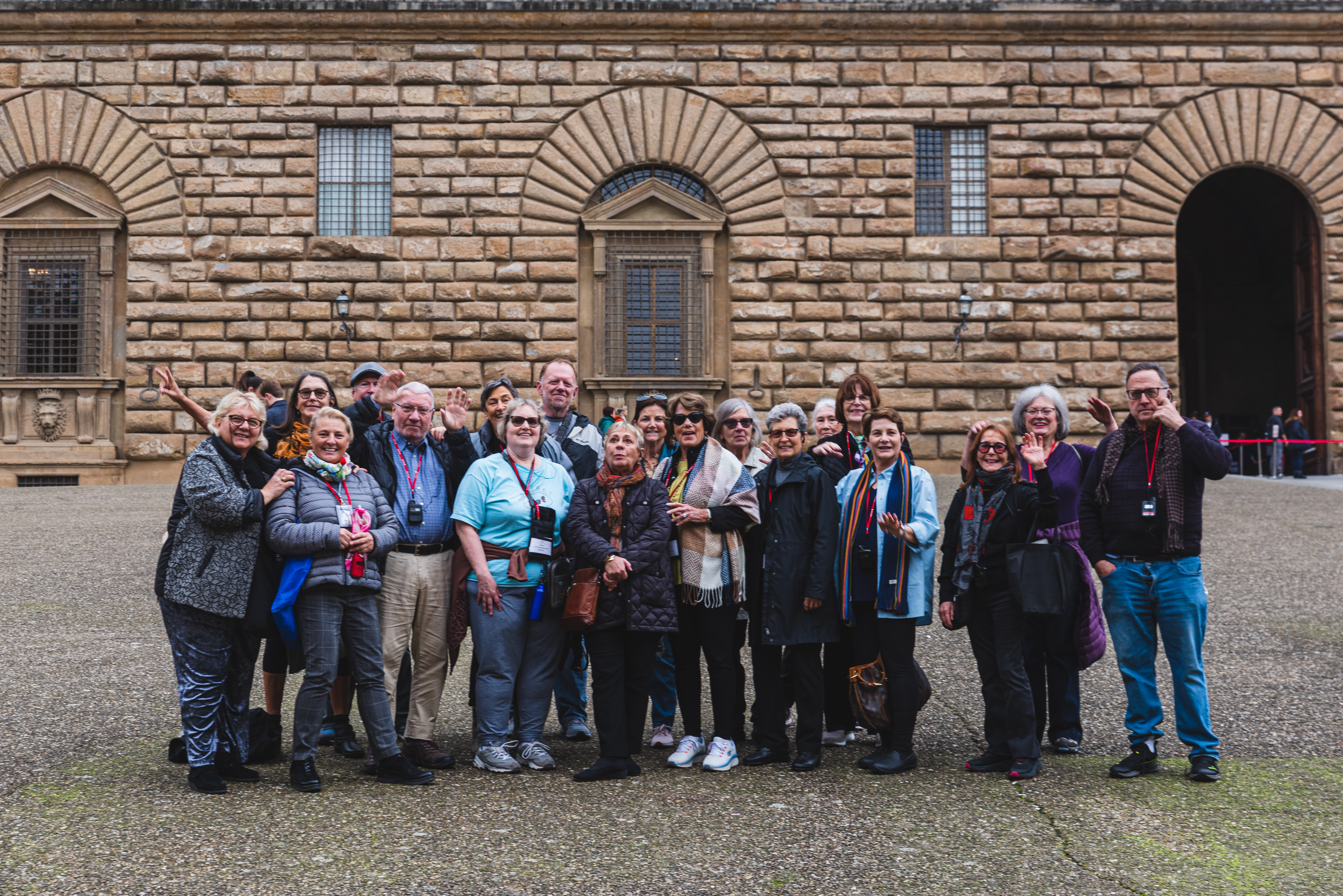 A tour group in Italy poses happily for a photo in the courtyard of a large, rusticated stone building, smiling and waving.