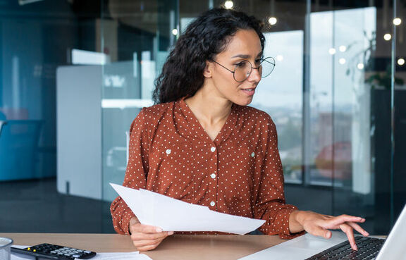 Business professional woman sitting at a desk looking at a laptop and document