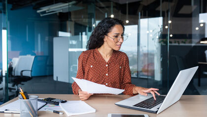 Business professional woman sitting at a desk looking at a laptop and document