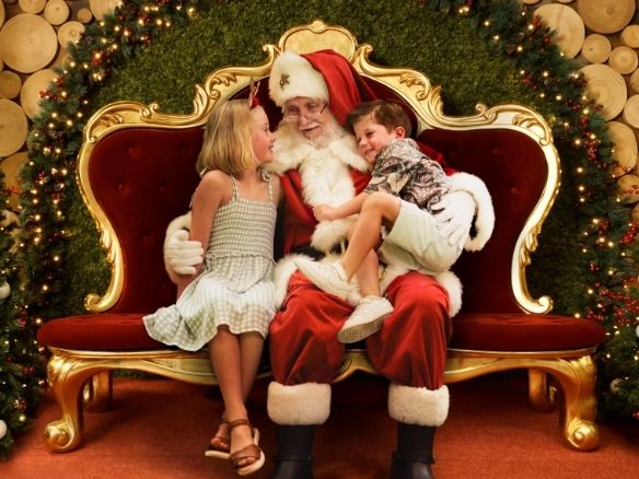 Two children sit with Santa on his red velvet throne, surrounded by lit Christmas garlands.