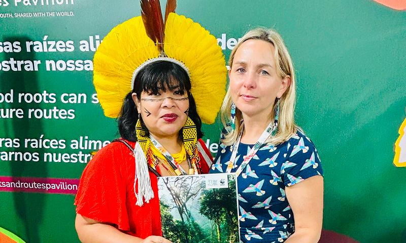 Two women sanding together in front of a sign holding a brochure, one of them is dressed in traditional aboriginal clothes and has face decorations