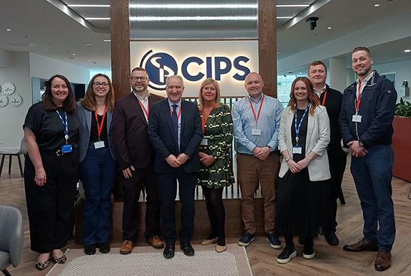 A group of eight people standing in a row in front of a wood-paneled wall with a glowing "CIPS" logo.
