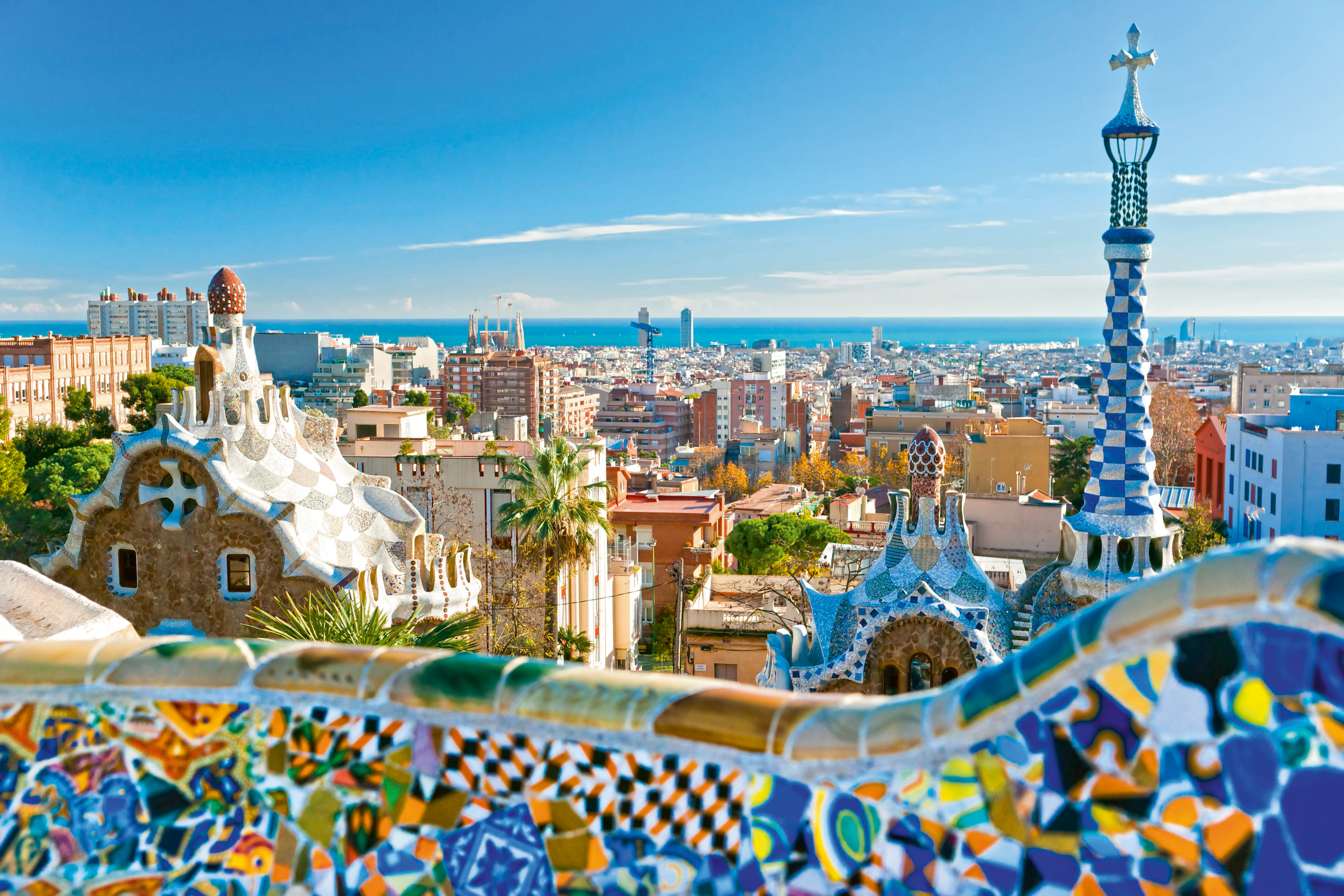 Colorful mosaic buildings at Park Guell overlook the Barcelona city skyline and the Mediterranean Sea under a clear blue sky.