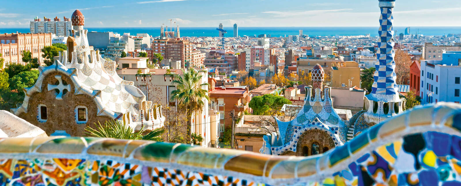 Colorful mosaic buildings at Park Guell overlook the Barcelona city skyline and the Mediterranean Sea under a clear blue sky.