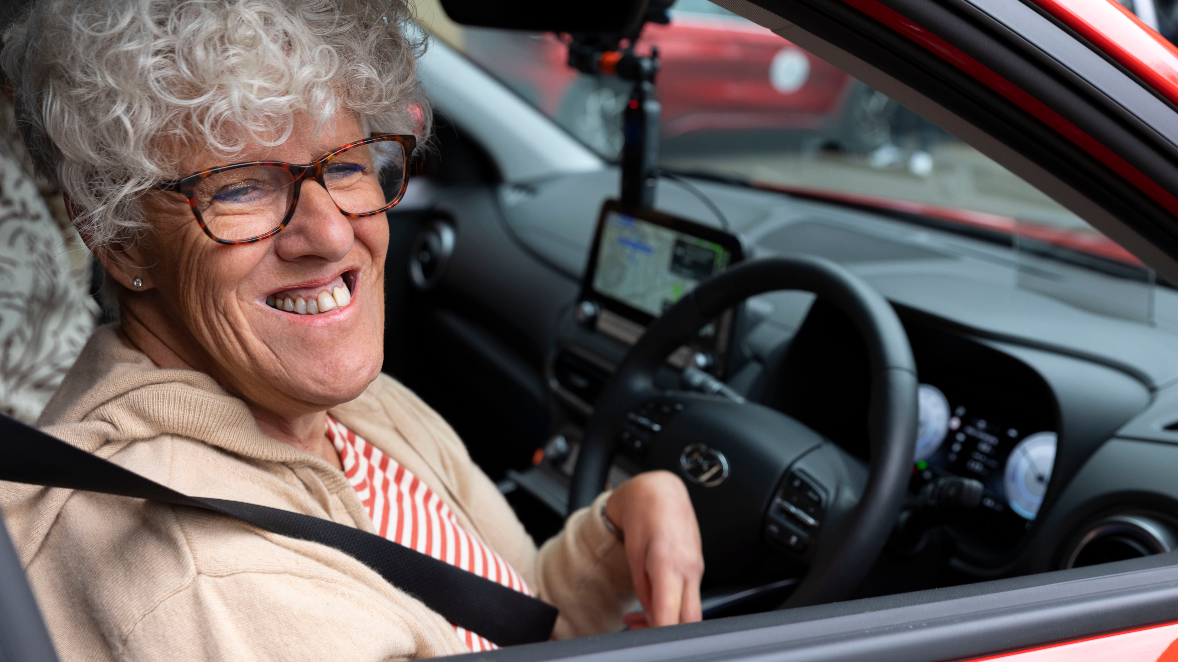 An older woman with curly hair and glasses smiling while sitting in the driver’s seat of a red car.