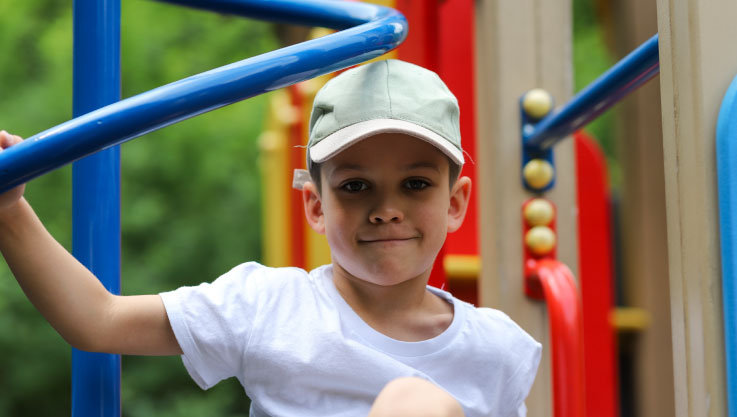 A close-up of a young boy wearing a white t-shirt and a light green baseball cap. He is looking directly at the camera with a slight smile while playing on a colorful outdoor playground structure. The background shows blurred green foliage.