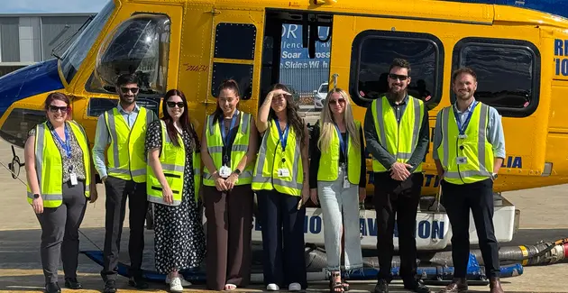 A group of eight people, a mix of men and women, stand in a line in front of a large, bright yellow and blue helicopter. All the individuals are wearing high-visibility yellow safety vests over their attire. The helicopter's open door is visible, and some text is legible on its side, including the partial words 