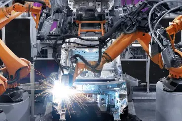 An image of an automated factory line with three large, orange industrial robotic arms assembling a car chassis. Bright sparks fly from a welding point in the center of the frame.