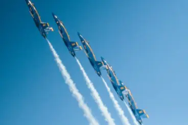 Four blue and yellow military fighter jets flying in a tight diagonal formation, leaving white contrails behind them against a clear blue sky.