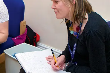 A close-up photo of a woman with blonde hair, wearing a dark top, a colorful beaded necklace, and a lanyard, writing in a large open notebook with a pen.