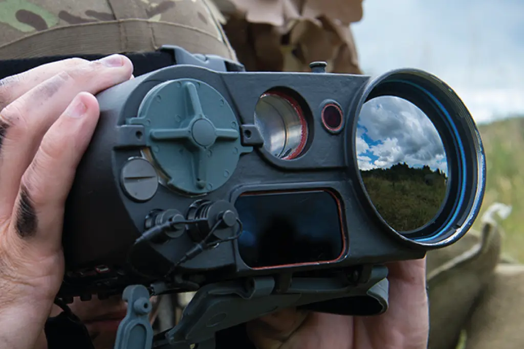 A close-up view of a person wearing a camouflage helmet and holding high-tech, dark gray military or professional binoculars, with a reflection of a cloudy sky and grassy field visible in the large front lens.