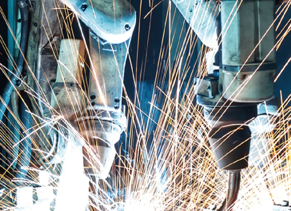 A close-up, dramatic shot of two industrial robotic welding arms in a dark setting, generating a bright shower of orange and white sparks during the welding process.