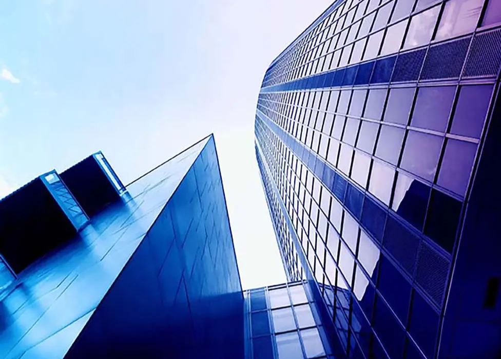 An abstract, low-angle shot of two adjacent modern skyscrapers against a bright sky. The buildings are primarily composed of reflective glass panels and appear in stark, cool colors of deep blue and purple due to color grading. The image emphasizes the sharp, angular geometry and verticality of the architecture.