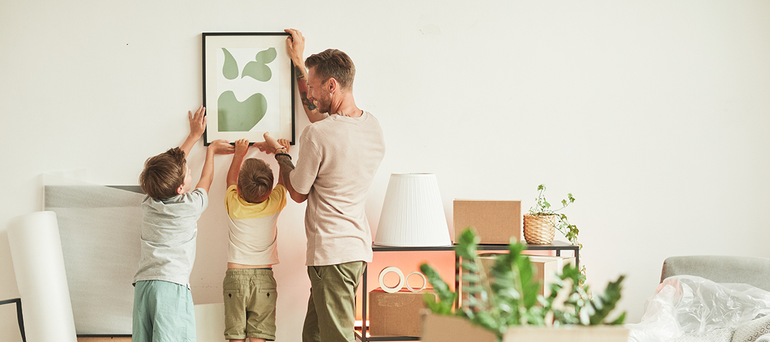 A white man hangs an print on the wall of room with help of two little kids