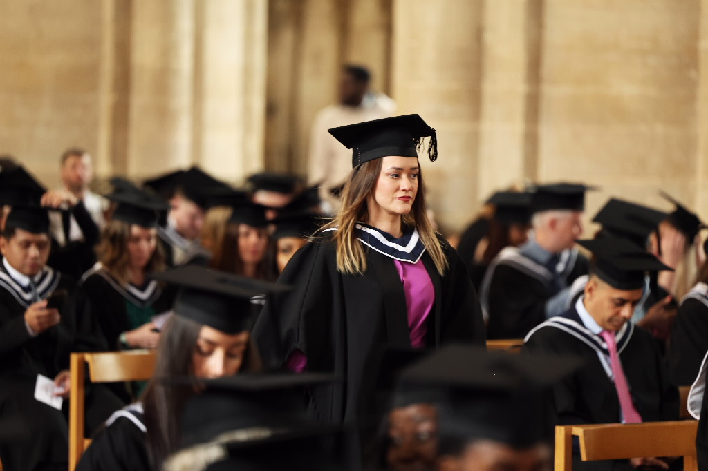 Female graduate standing in a purple top among rows of seated graduates.