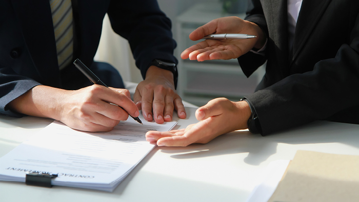 Two business people signing a contract. In view are their hands and a paper contract, with one person holding a pen over it as if they are about to sign.