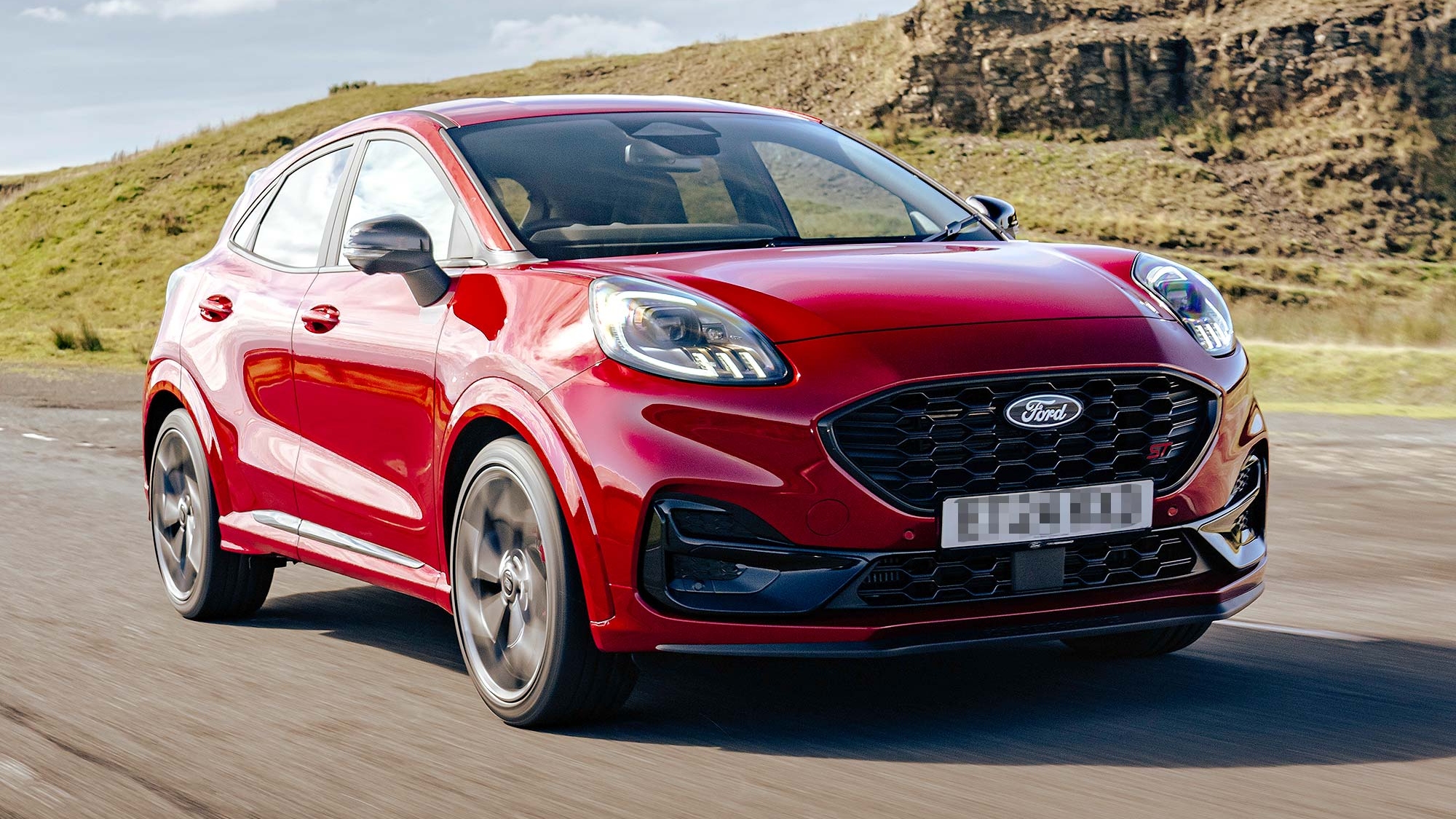 A red Ford Puma compact SUV driving on an open road with hills in the background, available on the Motability Scheme.