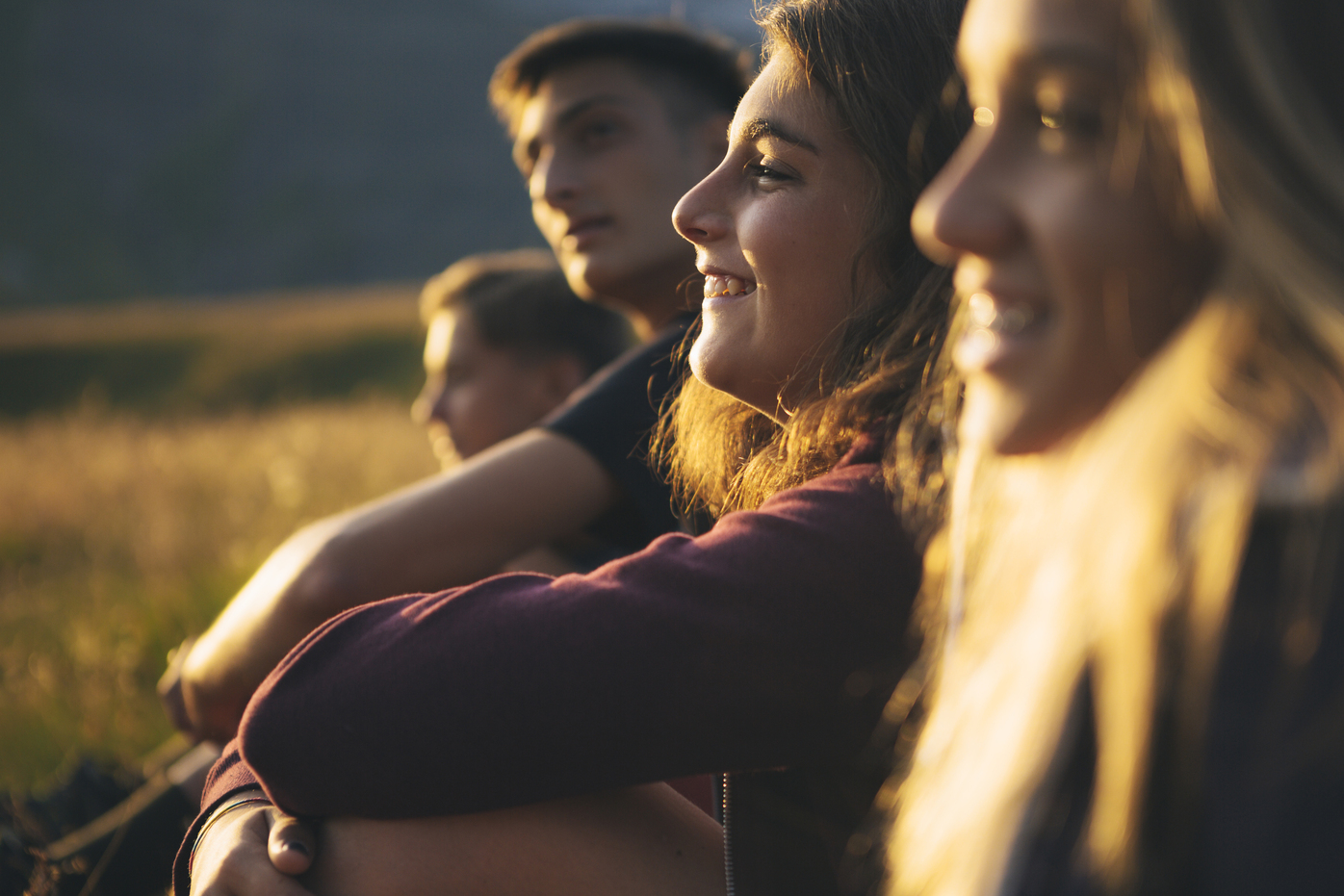 Four young friends sitting in a grassy field at sunset, smiling and enjoying the warm evening light.