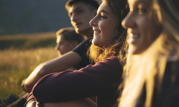 Four young friends sitting in a grassy field at sunset, smiling and enjoying the warm evening light.