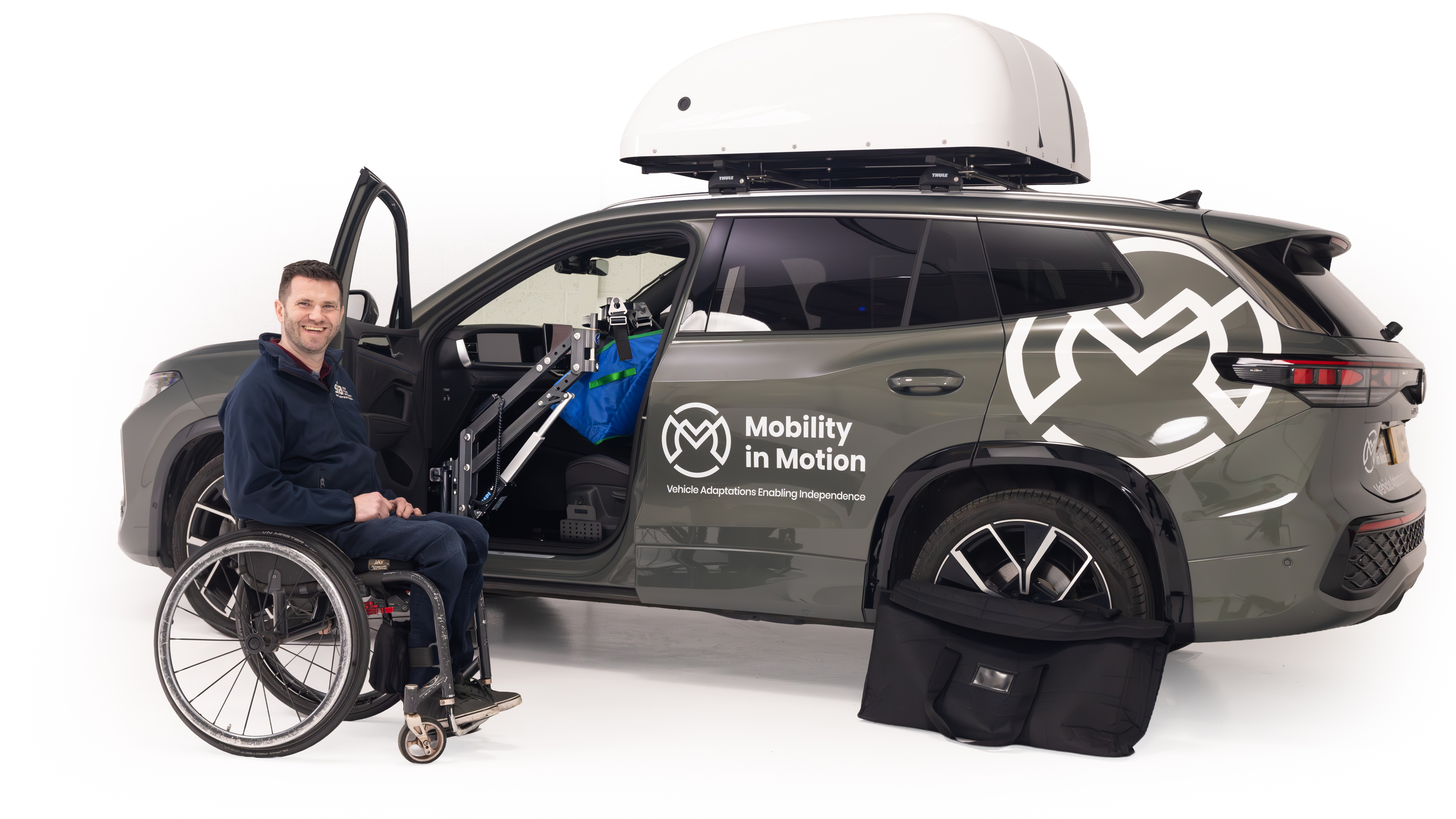 A wheelchair user beside a Mobility in Motion demonstration car fitted with a powered hoist to help him transfer into the passenger seat.