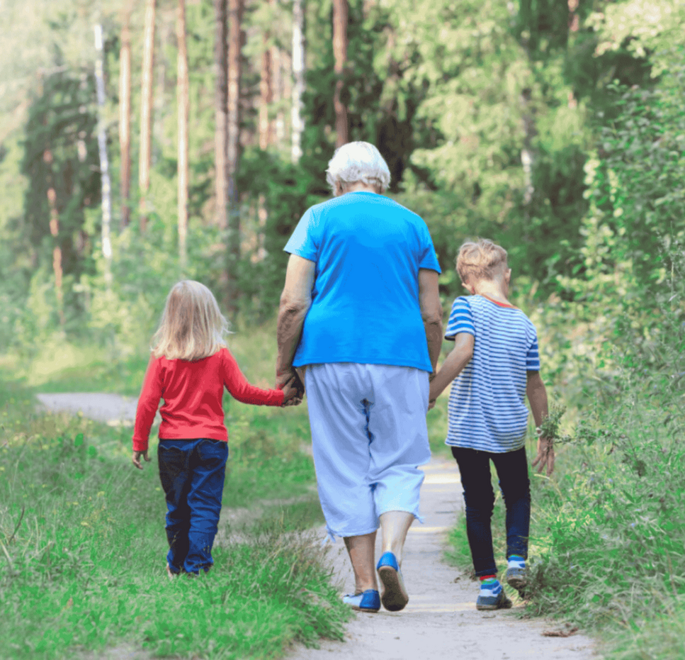 Older woman in an aqua blouse walks hand in hand with two young children through a forest, one wearing a bright red shirt and the other in blue stripes.