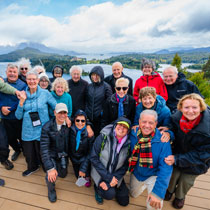 A group of travelers poses for a photo on a wooden deck overlooking a scenic lake and mountain landscape in Bariloche.