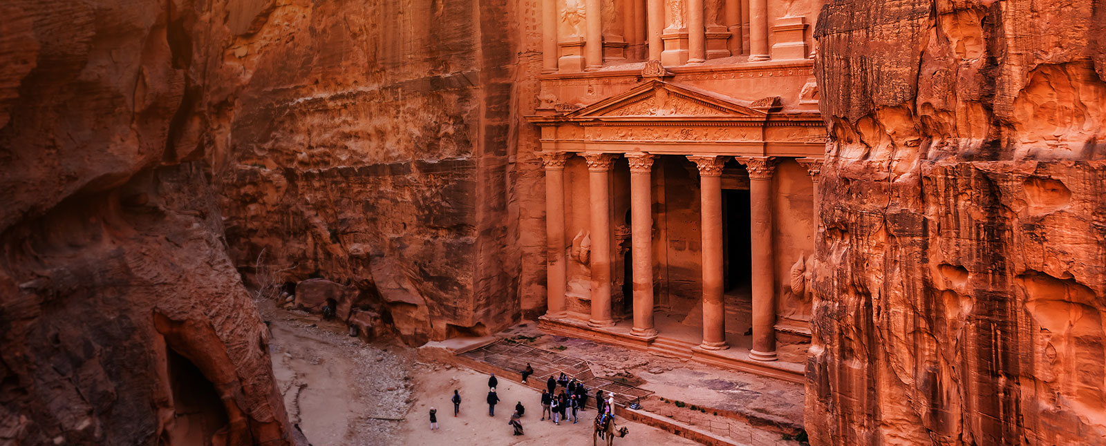 An elevated view of the Treasury at Petra, a historic facade carved into a red rock canyon, with tourists gathered on the ground below.