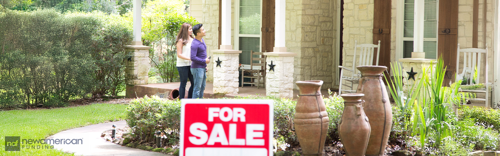 A white woman and an Asian man stand together in front of a large white stone home with a 