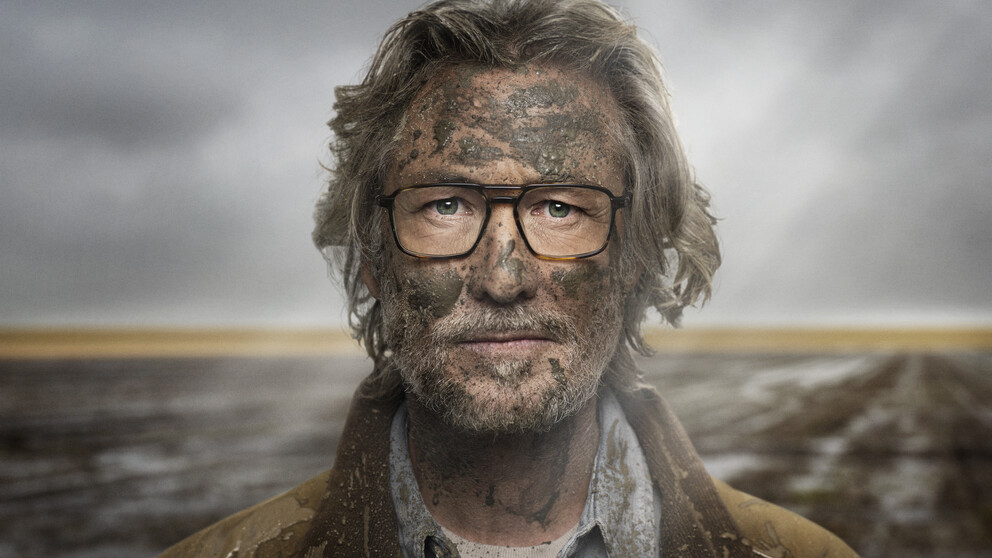 Older man standing outside in front of a muddy field, his face covered in mud but his glasses are clean