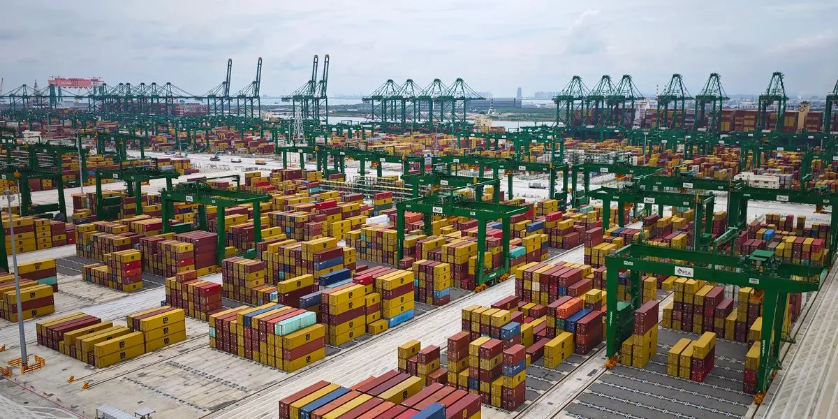 A large shipping port filled with stacks of colorful cargo containers and multiple green container cranes, with the sea and distant city skyline in the background.