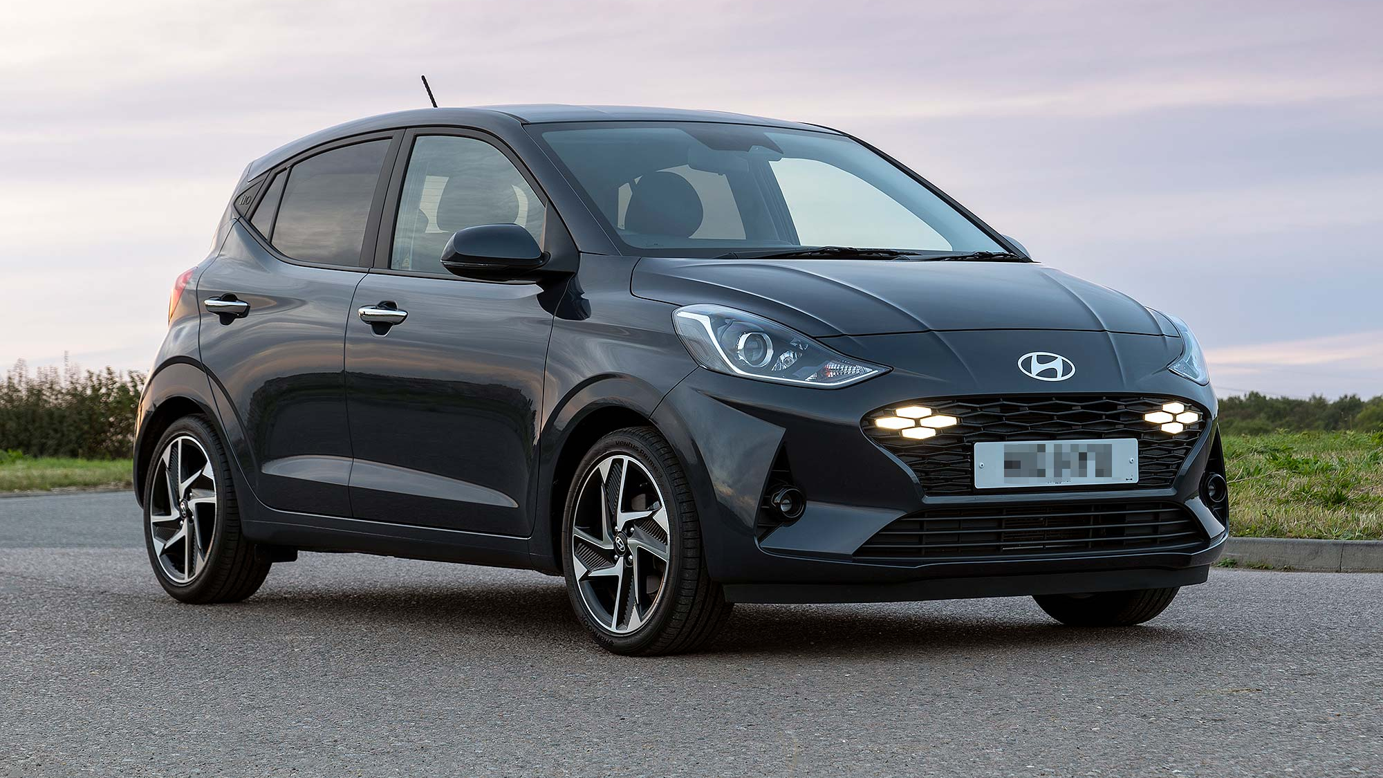 A dark grey Hyundai i10 city car is parked on a quiet road at dusk, with soft sky tones in the background.