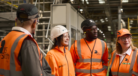 A diverse group of packaging plant employees are standing in a semicircle and looking at someone outside of the shot. They are all wearing safety equipment branded International Paper and DS Smith.