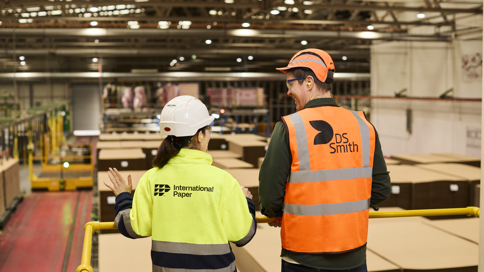 Two packaging plant workers, a man and a woman, seen from behind as they look over the plant. They are wearing reflective safety vests branded International Paper and DS Smith.
