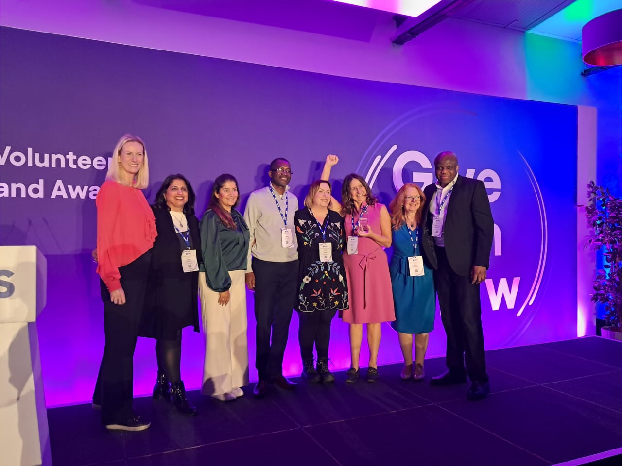 A group of eight people, four men and four women, standing together on a stage, posing for a photo. They are under purple lighting in front of a blue background with partial text visible for 'Volunteer Conference and Awards' and 'Give Gain Grow.' The woman in the pink dress is holding an award.