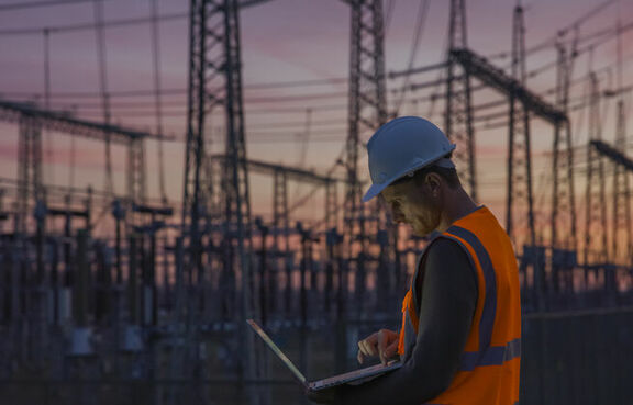 Man in safety vest and hard hat standing in front of a power station looking at a laptop