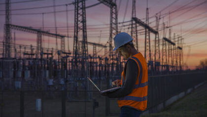 Man in safety vest and hard hat standing in front of a power station looking at a laptop