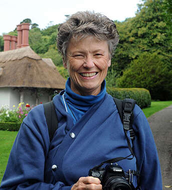 A smiling woman holding a camera stands on a path in front of a traditional thatched-roof cottage and garden in Ireland.