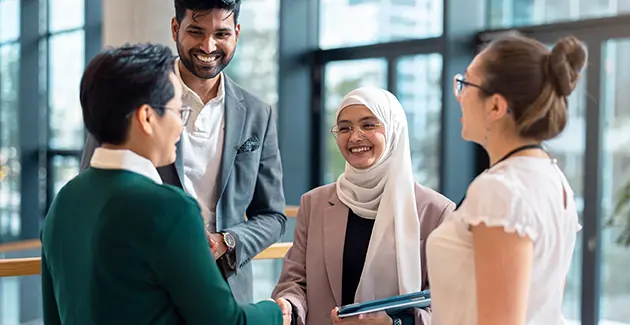 4 people smiling at each other with a woman in a hijab and pink blazer shaking hands with a man in a shirt and green jumper. 